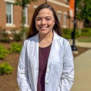 Dr. Jenna Bloemer standing outside of the college of pharmacy in her white coat