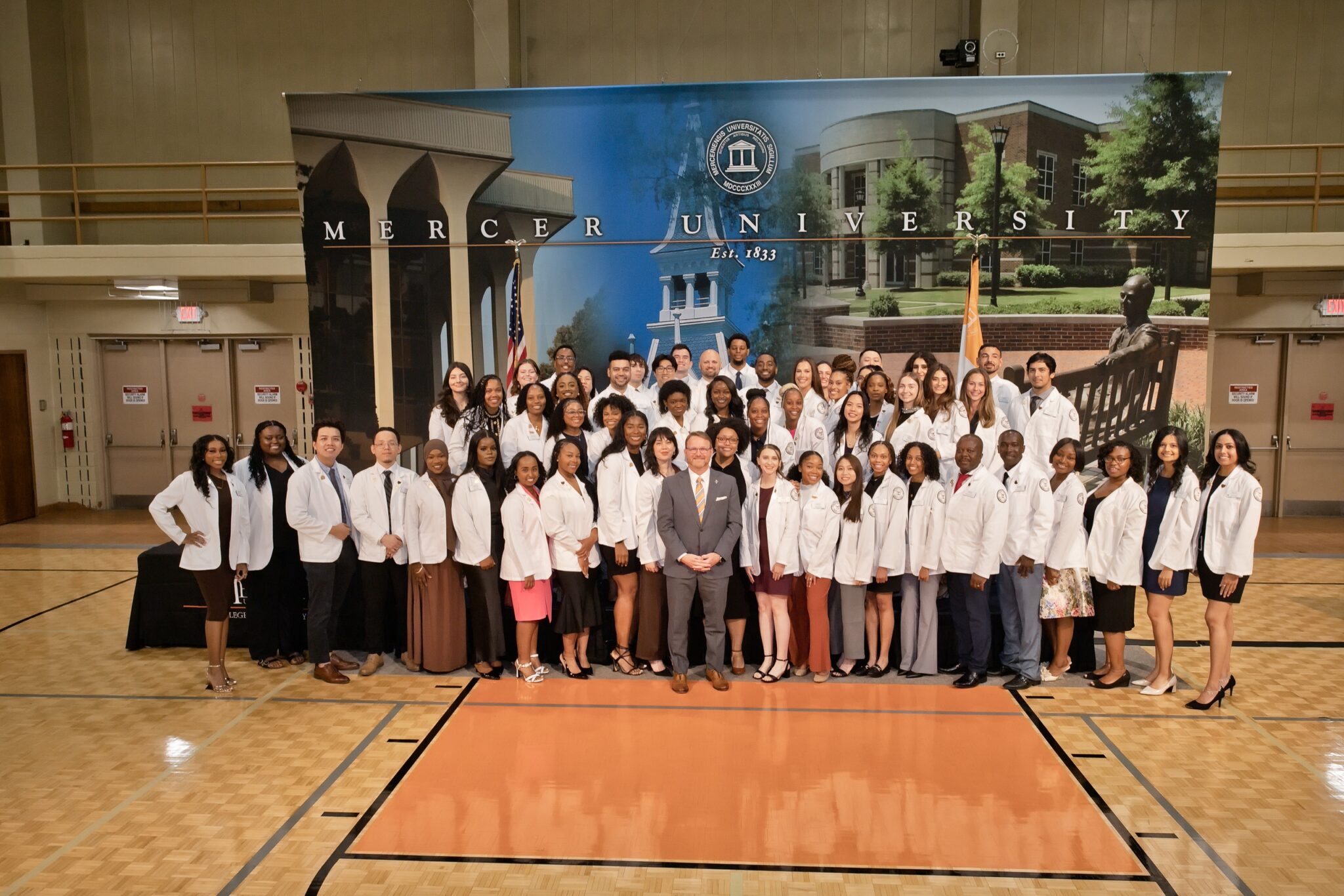 A group photo of the class of 2027 during their pinning ceremony in a gym. The background is a printed image of Mercer University