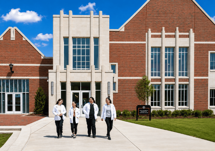 4 students walking outside the Tony and Nancy Moye Pharmacy and Health Sciences Center. The sky is blue.