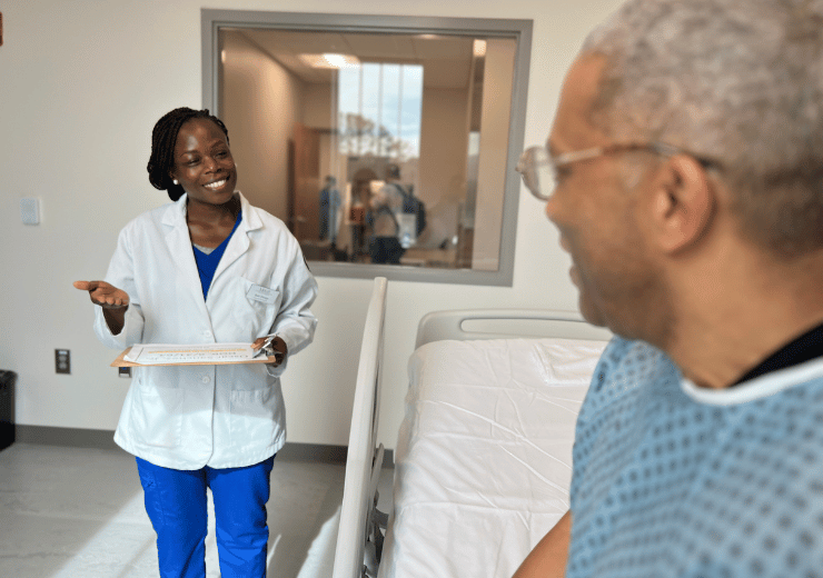 A student talking to a patient in Mercer's simulation lab. She is wearing a white coat while the patient is wearing a green hospital gown