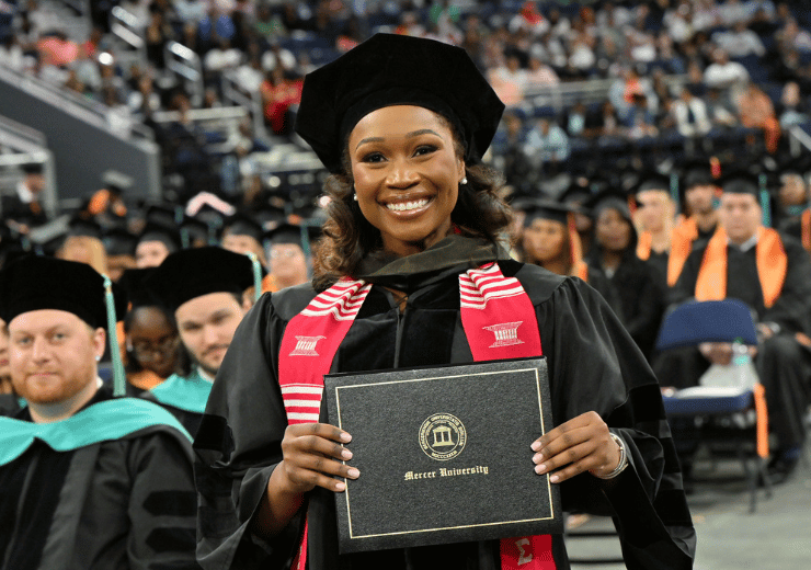 A woman in a red scarf and academic regalia smiles with her diploma in front of a large crowd