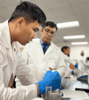 A student in a white lab coat mixing a solution in a compounding lab as a professor looks on
