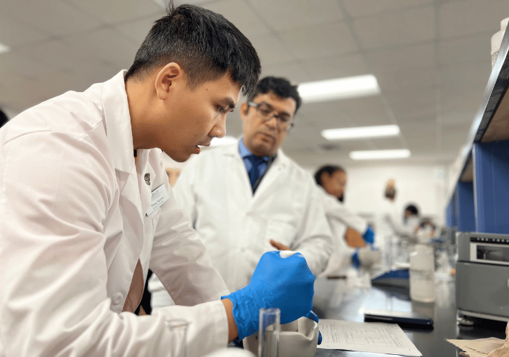 A student in a white lab coat mixing a solution in a compounding lab as a professor looks on
